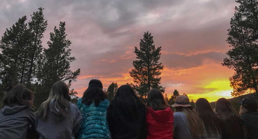 A group of people sit in a line, face away from the camera and watch the sun set. 